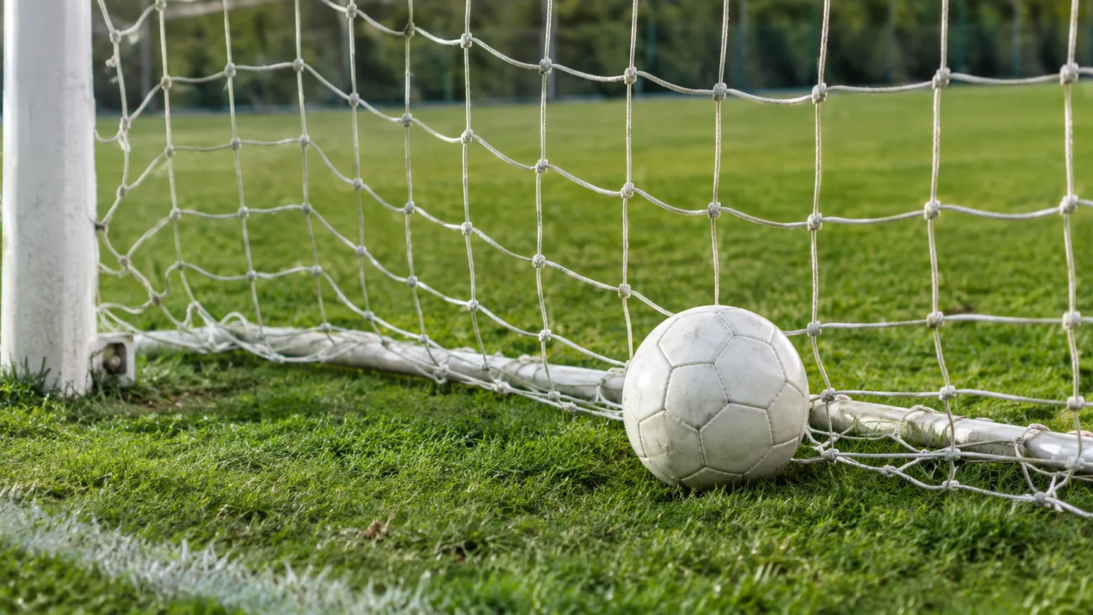 Ein Fußball liegt im Netz eines Tores auf einem leeren Rasenplatz.